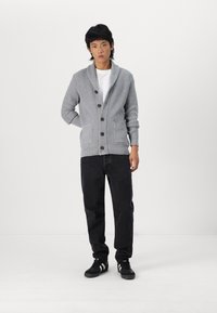 Grå strikket cardigan med sjalkrage, knappelukking og frontlommer, kombinert med svarte jeans og svarte joggesko.