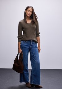 Olivengrüner gerippter Strickpullover mit V-Ausschnitt, kombiniert mit weit geschnittenen blauen Jeans. Dunkelbraune Wildleder-Handtasche und kurze braune Stiefel.