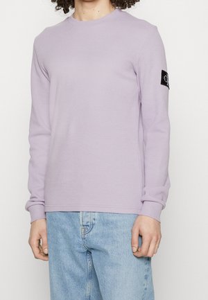 Person trägt einen langärmeligen hellvioletten Pullover mit einem schwarzen Patch am linken Arm und hellblauen Jeans, steht vor weißem Hintergrund.