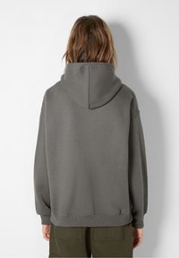 Persona con capelli castani alla spalla che indossa un ampio maglione grigio con cappuccio e pantaloni verdi, volto all'indietro su uno sfondo bianco semplice.