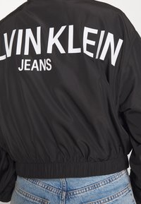 Černá bomber bunda s zipovým zapínáním, elastickým pasem a velkým bílým logem "CALVIN KLEIN JEANS" na zádech. Hladká textura.