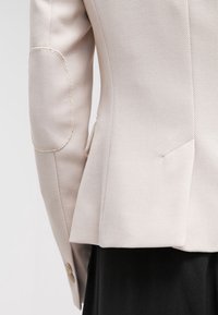 Gros plan sur la manche d'un blazer beige texturé avec un renfort au coude et des boutons, porté par-dessus un vêtement noir.