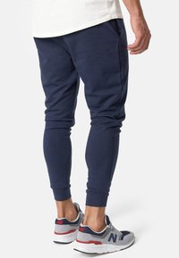 Marineblaue Jogginghosen aus weichem Stoff, mit schmal zulaufendem Bein und gerippten Bündchen. Kombiniert mit grau-rote Sneakers und einem weißen Oberteil.