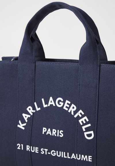 Морска канвас чанта с двоен дръжки, с бял печатен текст, гласящ "KARL LAGERFELD PARIS 21 RUE ST-GUILLAUME." Гладка текстура.