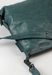 Borsa in pelle teal con una texture liscia, dotata di chiusura con zip e tracolla rimovibile impresso con il logo “aunts & uncles”.