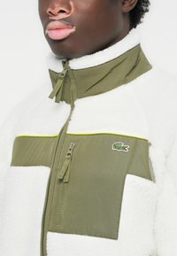 Oliven grønn fleecejakke med hvite panele av pels, en med glidelås lomme og neongul kant. Har Lacoste-logo på brystet.