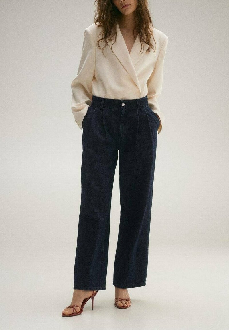 Femme portant un blazer crème, un pantalon large taille haute foncé et des sandales marron à talons avec des brides, debout les mains dans les poches.