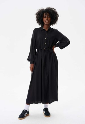Une robe chemise noire avec un cordon de serrage à la taille, des manches longues et une silhouette fluide, assortie à des baskets noires et des chaussettes blanches.