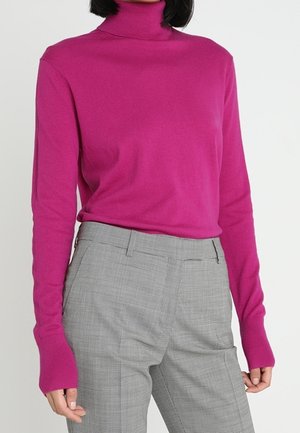 Woman wearing a bright pink turtleneck sweater tucked into high-waisted gray tailored trousers against a plain background.