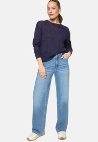 Femme portant un pull bleu marine, un jean large bleu clair et des baskets blanches, debout avec une main dans la poche devant un fond uni.