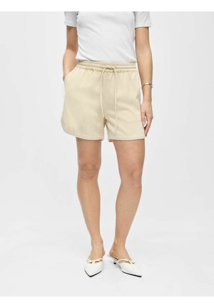 Mujer con shorts beige de cintura elástica, camiseta blanca de manga corta y mules blancos de tacón con punta puntiaguda, de pie con las manos en los bolsillos.