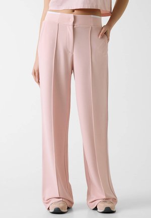 Femme portant un pantalon large texturé rose pâle taille haute avec plis devant et des baskets assorties, main dans la poche.