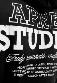 Schwarze Kapuzenjacke mit großen weißen Schriftzug "APREL STUDIOS" und kleinerem weißen Text darunter mit verschiedenen Aussagen zur Marke.