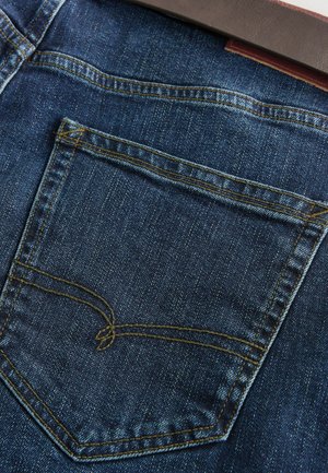 Donkerblauwe spijkerbroek van denim met een enkele achterzak met gele stiksels. De textuur is glad met een lichte glans.