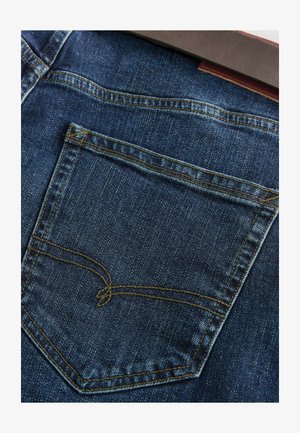 Tamsiai mėlynos džinsinės kelnės, turinčios vieną galinį kišenę su geltonomis siūlėmis. Paviršius lygus, su švelniu blizgesiu.