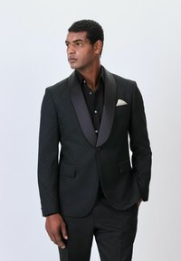 Must musta ülikond, millel on satiinist nurgelised kraed, struktureeritud kangas, millel on teemantkujuline muster, ühe nööbi kinnitamine ja valge taskurätt.