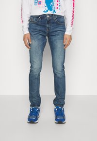 Siniset denim-farkut kapea leikkaus, jossa on klassinen viiden taskun muotoilu ja hienovarainen kuluminen. Yhdistetty sinisiin urheilukenkään.