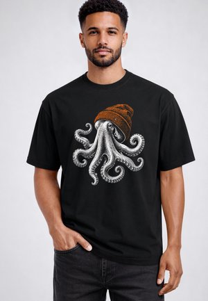 Mann trägt ein schwarzes T-Shirt mit einem detaillierten weißen Oktopus-Design, der eine braune Mütze trägt und mit einer Hand in der Tasche steht.