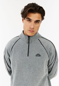 Grauer Fleece-Pullover mit einem Halbreißverschluss-Design, schwarzen Akzenten und einem kleinen Logo auf der Brust. Sanfte Textur und entspannte Passform.