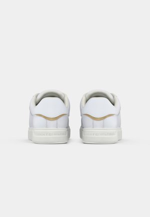 Para niskich białych sneakersów widzianych od tyłu, z beżowym akcentem nad piętą i białymi podeszwami z logo.