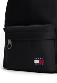 Mochila de nylon negra con un bolsillo frontal con cremallera, herrajes plateados y un parche con el logo "TOMMY JEANS" en rojo, blanco y azul.