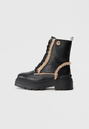 Botas de tornozelo em couro preto com sola grossa, apresentando forro felpudo em tom claro, ilhós dourados e detalhe do logotipo lateral. Fecho com atacadores.
