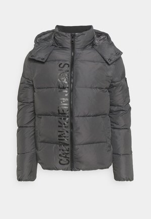 Veste d'hiver matelassée noire avec fermeture éclair frontale, deux poches, capuche amovible et logo vertical Calvin Klein Jeans sur le côté gauche devant.