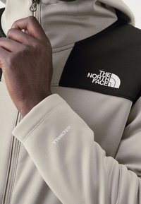 Gros plan d'une main tirant la fermeture éclair d'une veste beige et noire The North Face avec le texte WindWall sur la manche.