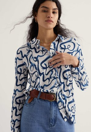 Frau trägt ein weißes Hemd mit blauen abstrakten Mustern, blaue Jeans, braunen Gürtel sowie goldene Creolen und Halsketten.