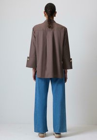 Camisa marrón de botones con mangas largas, combinada con jeans de mezclilla azul de pierna ancha. La tela parece suave con un diseño estructurado.