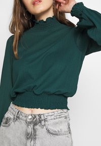 Långärmad, teal färgad kort topp med volangkrage och elastisk midja, kombinerat med högmidjade, ljusgrå denimjeans.