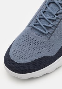 Zapatilla de malla azul claro y navy con parte superior texturizada, punta redonda y suela blanca plana. Presenta cordones trenzados y costuras reforzadas.