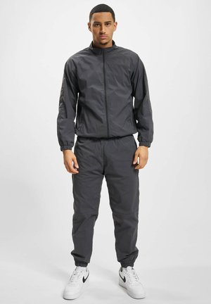 Mann steht in dunkelgrauer Zip-Up-Jacke und Hose mit weißen Sneakern vor schlichtem hellem Hintergrund.