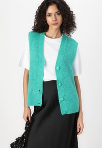 Turquoise knitted vest with a ribbed texture, featuring five buttons, worn over a white T-shirt, paired with a black skirt.