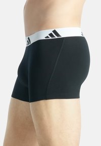 Boxer briefs pretos feitos de tecido elástico, com um cós elástico branco com riscas negras e logótipo, projetados para conforto e ajuste.