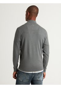 Pull gris à manches longues en tricot avec un col haut, des poignets et un ourlet côtelés. Porté par-dessus une chemise blanche, présentant des motifs texturés subtils.
