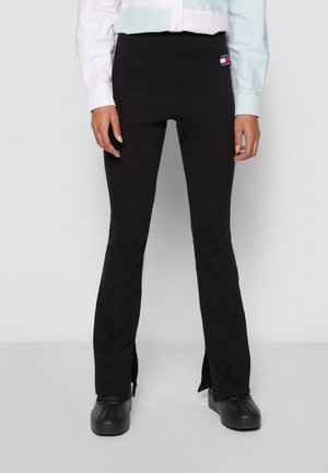 Vrouw draagt zwarte slim-fit broek met voorste splits aan de zoom, een lichtblauw-wit gestreept overhemd met knopen en zwarte plateauzolen.