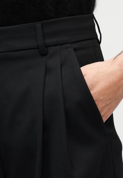 Μαύρο παντελόνι ραμμένο κατά παραγγελία με πιέτες στο μπροστινό μέρος, λεπτομέρεια με μπροστινές τσέπες και λείο, υφασμάτινο φινίρισμα με υφή.