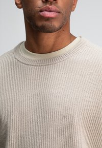 Beige gerippter Strickpullover mit rundem Ausschnitt, der einen helleren Innenkragen hat. Nahaufnahme betont die Textur und die Nähdetails.