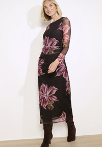 Žena v dlouhých černých šatech s velkými růžovými květinovými vzory a průhlednými rukávy, v kombinaci s tmavě hnědými vysokými botami na podpatku.