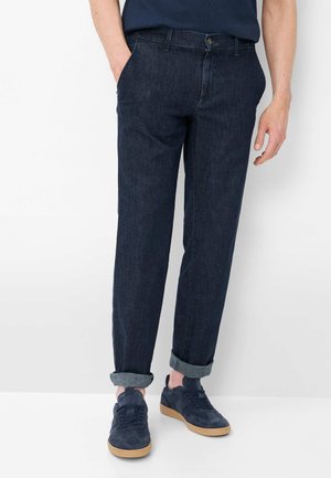 Mann trägt dunkle, hochgekrempelte Jeans und marineblaue Sneaker mit Gummisohlen, steht vor weißem Hintergrund.