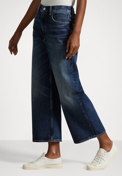 Πανάκριβα τζιν παντελόνια σε σκούρο μπλε απόχρωση με φαρδιά γραμμή και ψηλή μέση, που διαθέτουν κλείσιμο με κουμπί, τσέπες και διακριτική φθορά. Συνδυασμένα με ανοιχτόχρωμα αθλητικά παπούτσια.