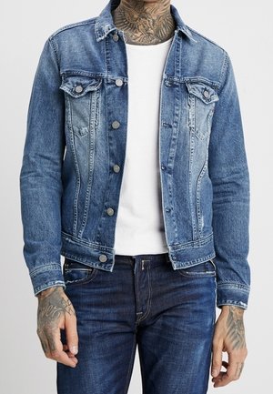 Denimjack in lichtblauw met een knoopsluiting aan de voorkant, twee borstzakken en een standaard kraag. Draagt over een eenvoudig wit T-shirt en donkere jeans.