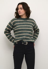 Pulover din lână cu dungi verzi și crem, cu guler rotunjit și textură groasă, asociat cu blugi negri și o curea cu cataramă argintie.