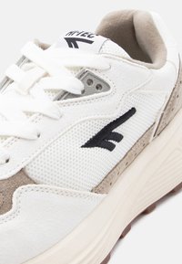 Κοντινή λήψη λευκού και μπεζ αθλητικού παπουτσιού με πάνελ από δίχτυ, παχύ πέλμα, λευκά κορδόνια και μαύρο κεντημένο λογότυπο στο πλάι.