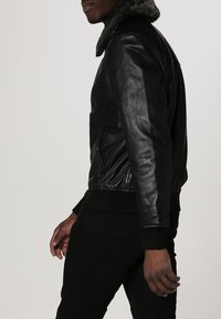 Černá kožená bunda s texturovaným, chlupatým límcem; má boční kapsy a žebrované manžety; přiléhavý střih zdůrazňuje siluetu nositele.