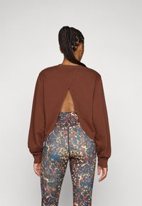 Sweatshirt marron avec un dos ouvert, associé à un legging multicolore à motifs comprenant des teintes de bleu, d'orange et de vert.
