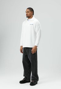 Witte fleece pullover met kraag en blauw geborduurde tekst, gecombineerd met donkergrijze losse jeans en zwarte chunky sneakers.