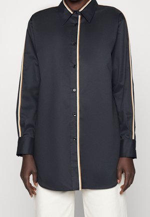 Chemise noire à manches longues boutonnée avec des rayures beiges et blanches au centre et sur les manches, portée avec un pantalon blanc.