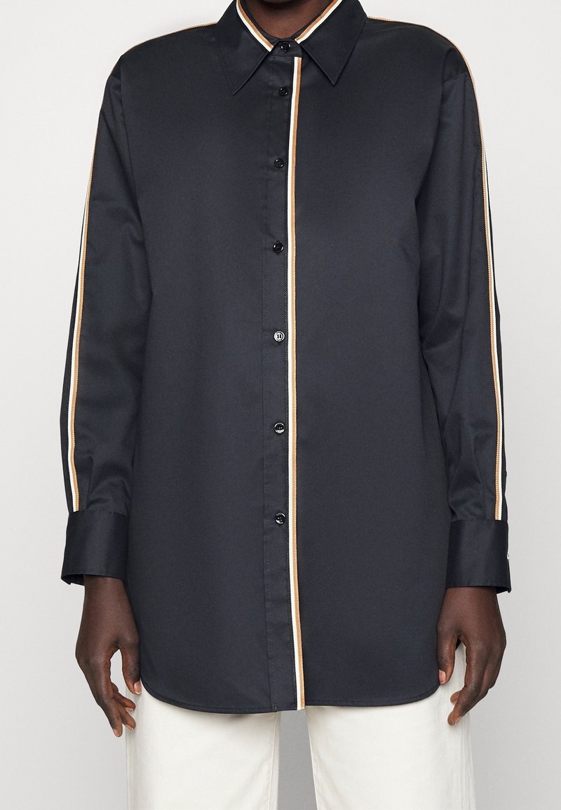 Chemise noire à manches longues boutonnée avec des rayures beiges et blanches au centre et sur les manches, portée avec un pantalon blanc.